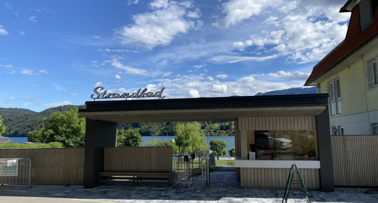Der neue Eingangsbereich des Strandbad Seeboden. Ein aus Holz gefertigtes kleines Gebäude mit der Aufschrift Strandbad. Zuätzlich gibt es ein Drehkreuz für die Kontrolle, nebenan ist der Kassenbereich hinter einen großen Glasscheibe.